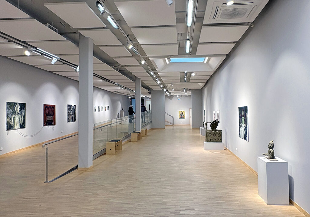 Rymlig konsthall med skulpturer och tavlor utställda längs väggarna. Takfönster och spotlights ger ett mjukt ljus över lokalen.