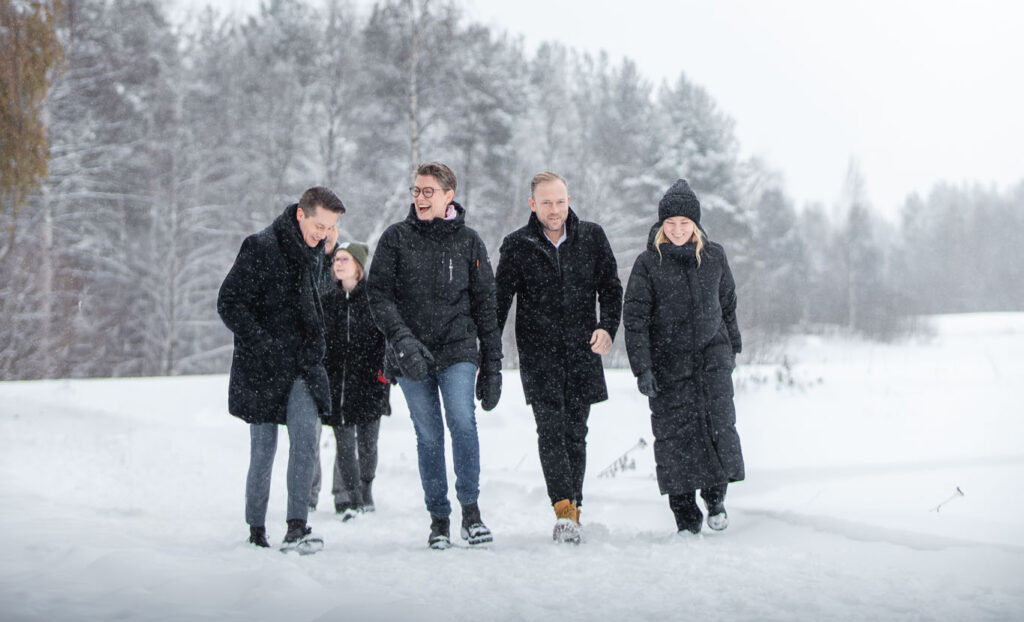 Fem personer promenerar tillsammans i ett snötäckt landskap. De bär mörka vinterkläder, snön faller och stämningen är lättsam och skrattfylld.