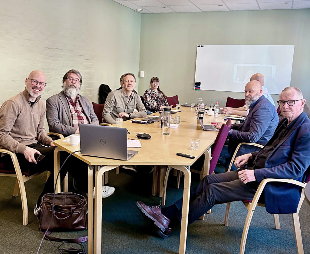 Personer sitter samlade runt ett konferensbord under ett styrelsemöte. På bordet står laptops, glas och kaffekoppar i ett ljust mötesrum med gröna väggar.