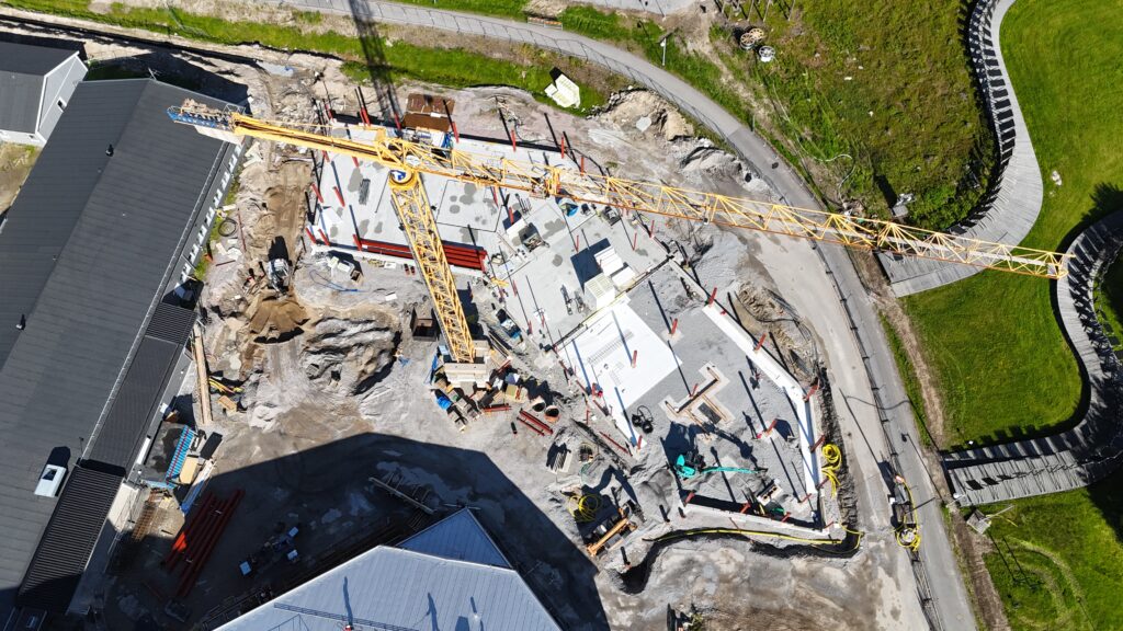 Flygbild över en byggarbetsplats där en stor betongplatta och röda stålpelare tar form. En tornkran sträcker sig tvärs över området.
