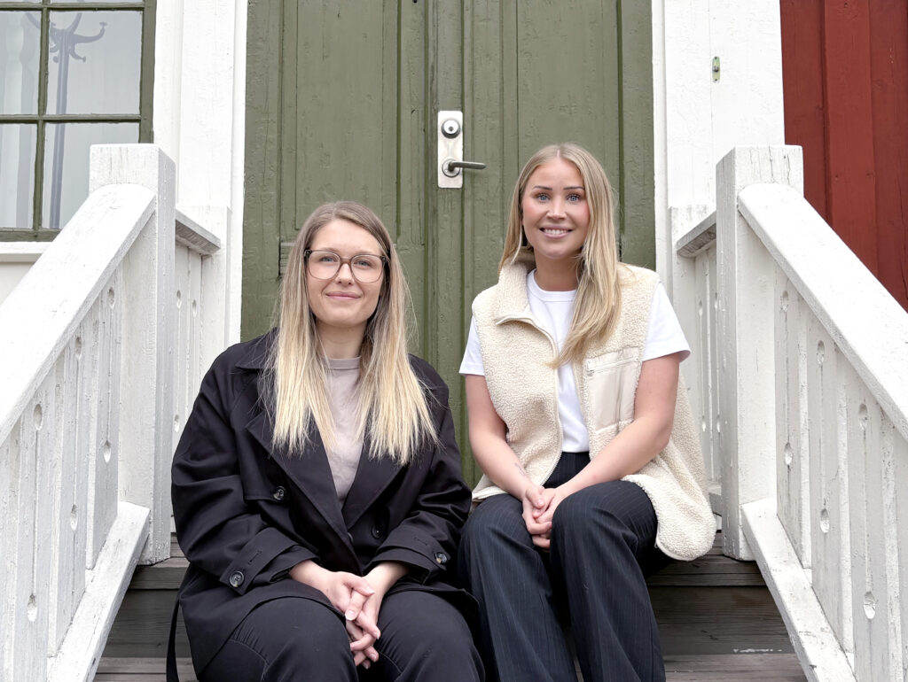 Två kvinnor sitter på en trätrappa framför en grön dörr på ett rött och vitt hus. De ler mot kameran i en lugn och välkomnande miljö.