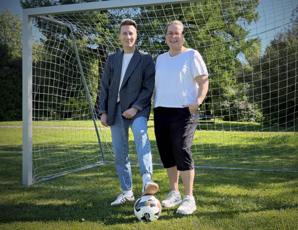 Två personer står framför ett fotbollsmål på en grön gräsmatta en solig dag. Personen till vänster har foten på en fotboll och bär mörk kavaj, ljusa jeans och vita sneakers. Personen till höger bär vit t-shirt, mörka shorts och vita sneakers. Båda ler mot kameran. I bakgrunden syns träd och grönska.