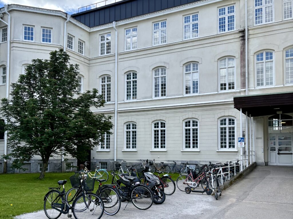 Vit byggnad i flera våningar med en grön innergård. Intill entrén står flera cyklar parkerade på en ny, ljusgrusad yta som omges av gräsmatta och träd.