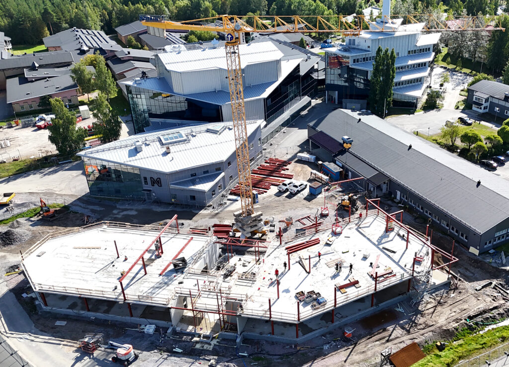 Drönarfoto över en pågående nyproduktion av ett kontorshus. Vädret är klart och soligt. Omgivningen visar ett flertalet fastigheter runt nybyggnationen, merparten av dessa har stora fönster- och glaspartier. En gul lyftkran är i centrum av bilden.