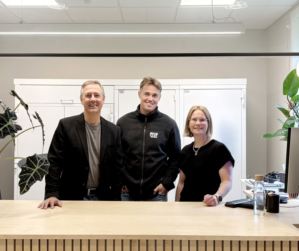 Tre personer står bakom en ljus träbänk på ett kontor. De ler mot kameran, omgivna av vita skåp, gröna växter och arbetsutrustning.