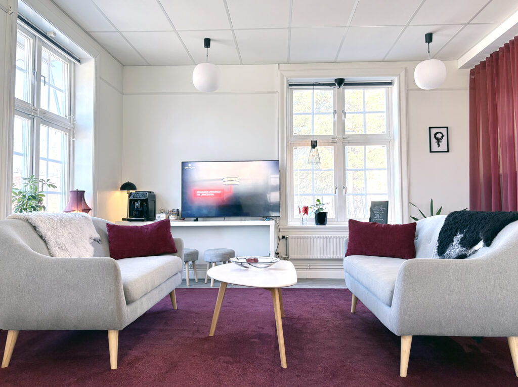 Interiörbild från hyresgäst. Vita väggar, grå soffor och flertalet vinröda inslag i form av draperi, kuddar och golvmatta. En stilren inredning. Höga fönster släpper in mycket dagsljus.