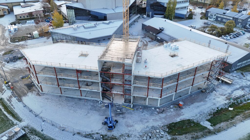 En flygbild visar en stor byggnad under uppförande, med flera våningsplan i betong och röda stålpelare. De stora, platta taken är täckta av ett ljust lager och omges av tillfälliga skyddsräcken. Mitt på byggnaden syns ett öppet trapphus med trappor mellan våningsplanen. En gul tornkran reser sig bakom byggnaden. På marken står en blå lift, och runt arbetsområdet finns byggmaterial, maskiner och tillfälliga stängsel. Ett tunt lager snö täcker marken och taken på de omgivande byggnaderna.