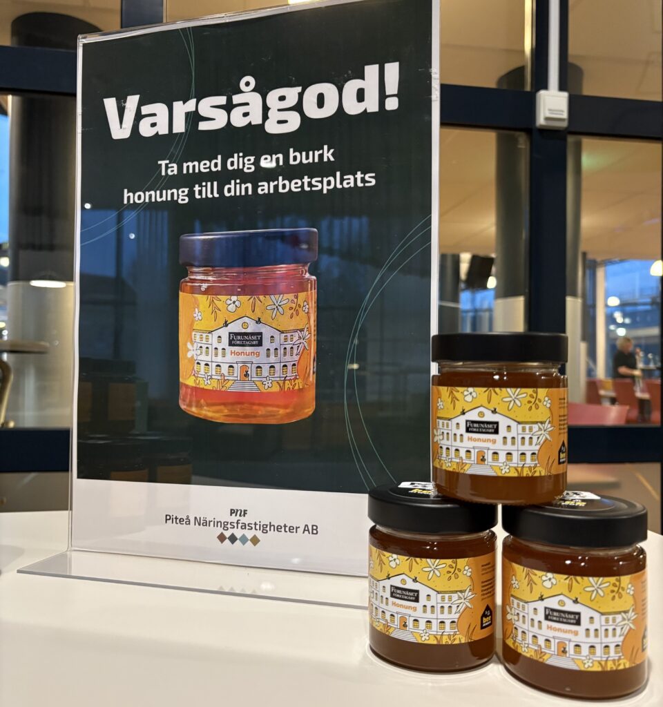 Tre honungsburkar med gula etiketter står framför en skylt med texten ”Varsågod! Ta med dig en burk honung till din arbetsplats”.
