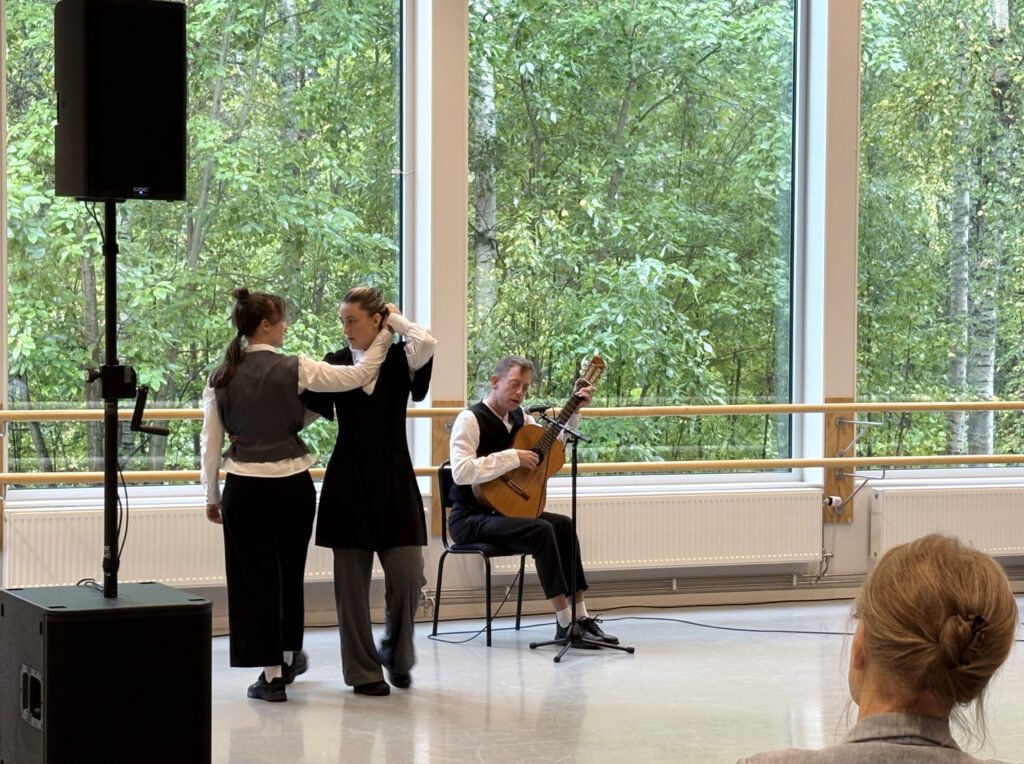 Två dansare möts i en rörelse framför stora fönster medan en sittande musiker spelar gitarr. Grönskande träd syns utanför.